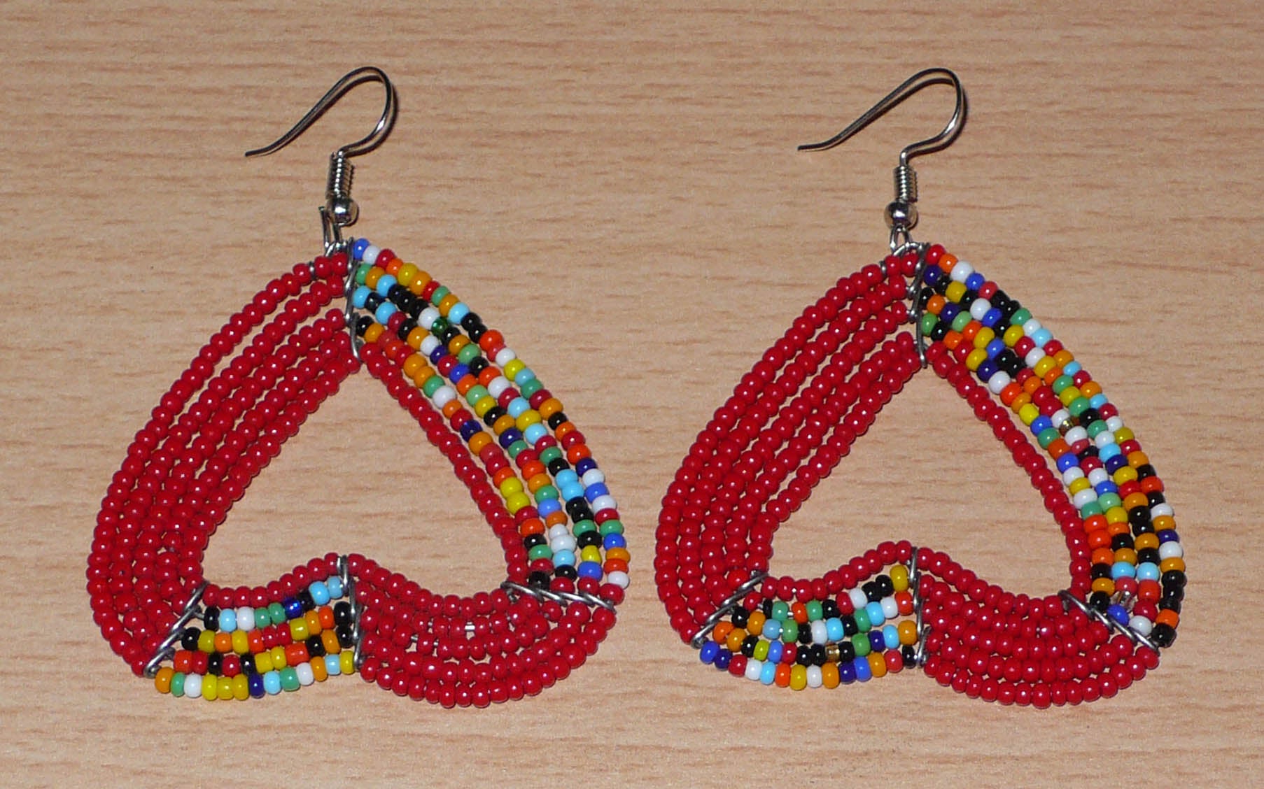 Boucles d'oreilles aux accents bohème composées de perles de rocaille rouges et d'un mélange de perles multicolores glissées sur cinq rangées de fils de fer montés en forme de cœurs inversés. De taille moyenne, elles mesurent 6,5 cm de long et 5 cm de large, et se portent avec des crochets en acier inoxydable sur des oreilles percées.