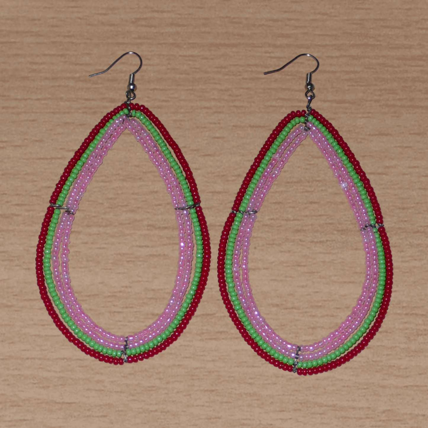 Boucles d'oreilles "oversized" composées de perles de rocaille rose nacré, vertes et rouges enfilées sur quatre rangées de fils de fer montés en forme de gouttes. Surdimensionnées, ces boucles d'oreilles mesurent 11 cm de long et 5,5 cm de large ; elles se portent avec des crochets en acier inoxydable sur des oreilles percées.