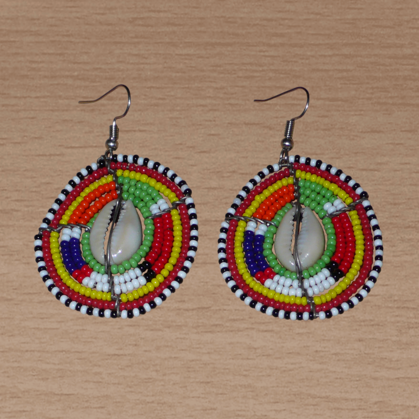 Boucles d'oreilles africaines ethniques aux motifs tribaux traditionnels caractéristiques de l'artisanat Massai. Confectionnées au Kenya, elles sont composées de perles de rocaille vertes et multicolores et de coquillages cauris fixés sur des fils de fer montés en forme de boucliers Massai. De taille moyenne, elles mesurent 6 cm de long et 4 cm de large, et se portent avec des crochets en acier inoxydable sur des oreilles percées.