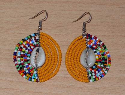 Boucles d'oreilles ethniques orange et multicolores fabriquées au Kenya composées de perles de rocaille et de coquillages cauris. De taille moyenne, elles mesurent 6 cm de long et 3,5 cm de large et se portent sur des oreilles percées.