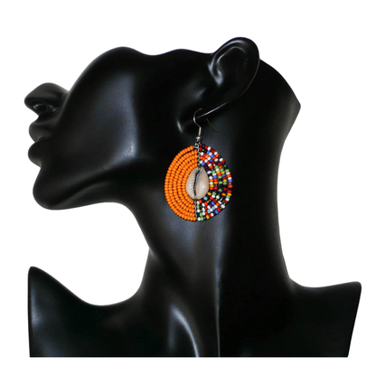 Boucles d'oreilles ethniques orange et multicolores fabriquées au Kenya composées de perles de rocaille et de coquillages cauris. De taille moyenne, elles mesurent 6 cm de long et 3,5 cm de large et se portent sur des oreilles percées.