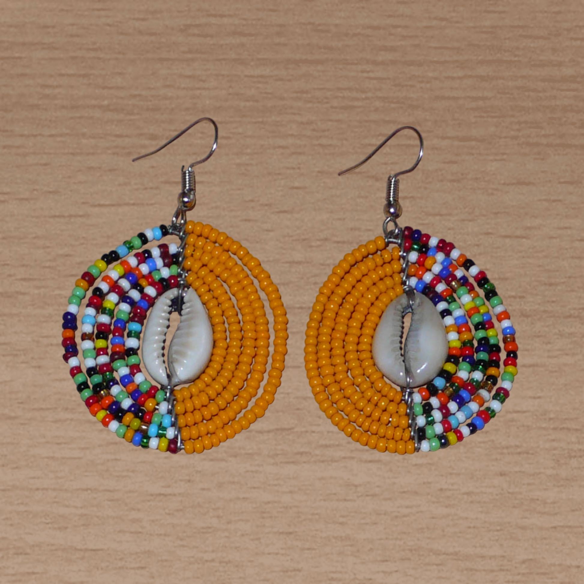 Boucles d'oreilles ethniques orange et multicolores fabriquées au Kenya composées de perles de rocaille et de coquillages cauris. De taille moyenne, elles mesurent 6 cm de long et 3,5 cm de large et se portent sur des oreilles percées.