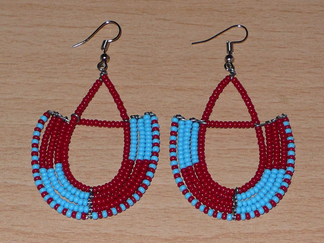 Boucles d'oreilles africaines Massai rouges et bleues composées de perles de rocaille glissées sur des structures géométriques en fils de fer montées en forme d'ancres. Ces boucles d'oreilles bicolores mesurent 7,5 cm de long et 4 cm de large et se portent avec des crochets en acier inoxydable. 