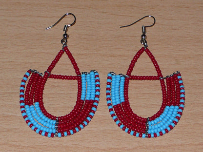 Boucles d'oreilles africaines Massai rouges et bleues composées de perles de rocaille glissées sur des structures géométriques en fils de fer montées en forme d'ancres. Ces boucles d'oreilles bicolores mesurent 7,5 cm de long et 4 cm de large et se portent avec des crochets en acier inoxydable. 