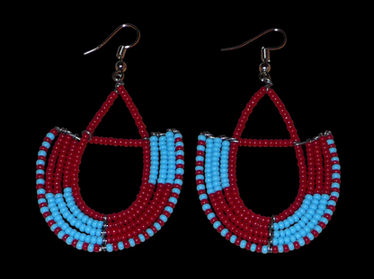Boucles d'oreilles africaines Massai rouges et bleues composées de perles de rocaille glissées sur des structures géométriques en fils de fer montées en forme d'ancres. Ces boucles d'oreilles bicolores mesurent 7,5 cm de long et 4 cm de large et se portent avec des crochets en acier inoxydable.
