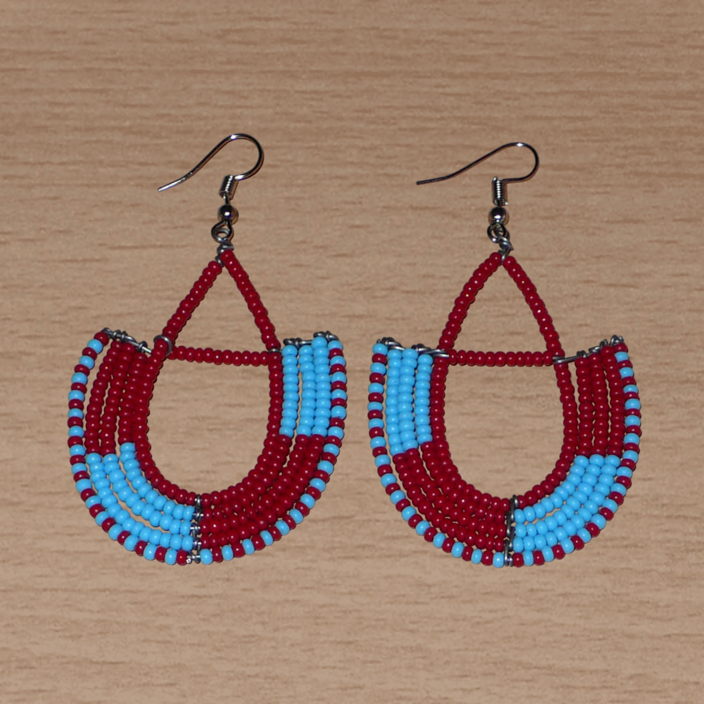 Boucles d'oreilles africaines Massai rouges et bleues composées de perles de rocaille glissées sur des structures géométriques en fils de fer montées en forme d'ancres. Ces boucles d'oreilles bicolores mesurent 7,5 cm de long et 4 cm de large et se portent avec des crochets en acier inoxydable.