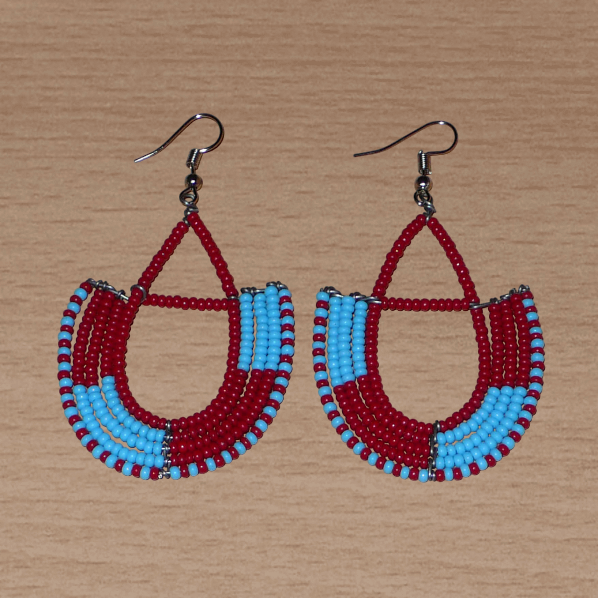 Boucles d'oreilles africaines Massai rouges et bleues composées de perles de rocaille glissées sur des structures géométriques en fils de fer montées en forme d'ancres. Ces boucles d'oreilles bicolores mesurent 7,5 cm de long et 4 cm de large et se portent avec des crochets en acier inoxydable.