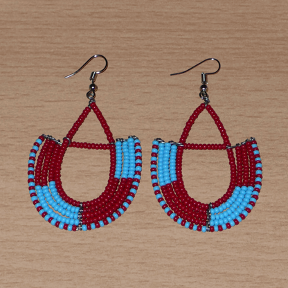 Boucles d'oreilles africaines Massai rouges et bleues composées de perles de rocaille glissées sur des structures géométriques en fils de fer montées en forme d'ancres. Ces boucles d'oreilles bicolores mesurent 7,5 cm de long et 4 cm de large et se portent avec des crochets en acier inoxydable.