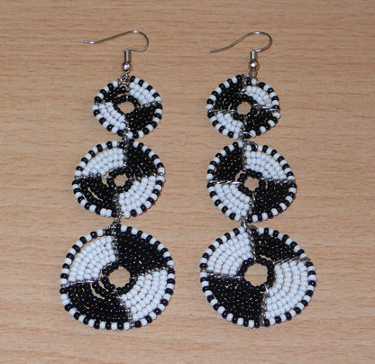 Longues boucles d'oreilles ethniques Massai composées de perles noires et blanches montées en cercles superposés. Ces boucles d'oreilles bicolores mesurent 9,5 cm de long et 3 cm de large ; elles se portent avec des crochets sur des oreilles percées.