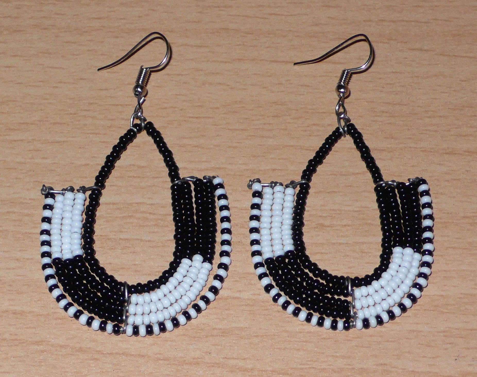 Boucles d'oreilles ethniques bicolores en forme d'ancres composées de perles de rocaille noires et blanches glissées sur des structures géométriques en fils de fer. De taille moyenne, ces boucles d'oreilles aux accents afro chic mesurent 7 cm de long et 4 cm de large ; elles se portent avec des crochets en acier inoxydable sur des oreilles percées.