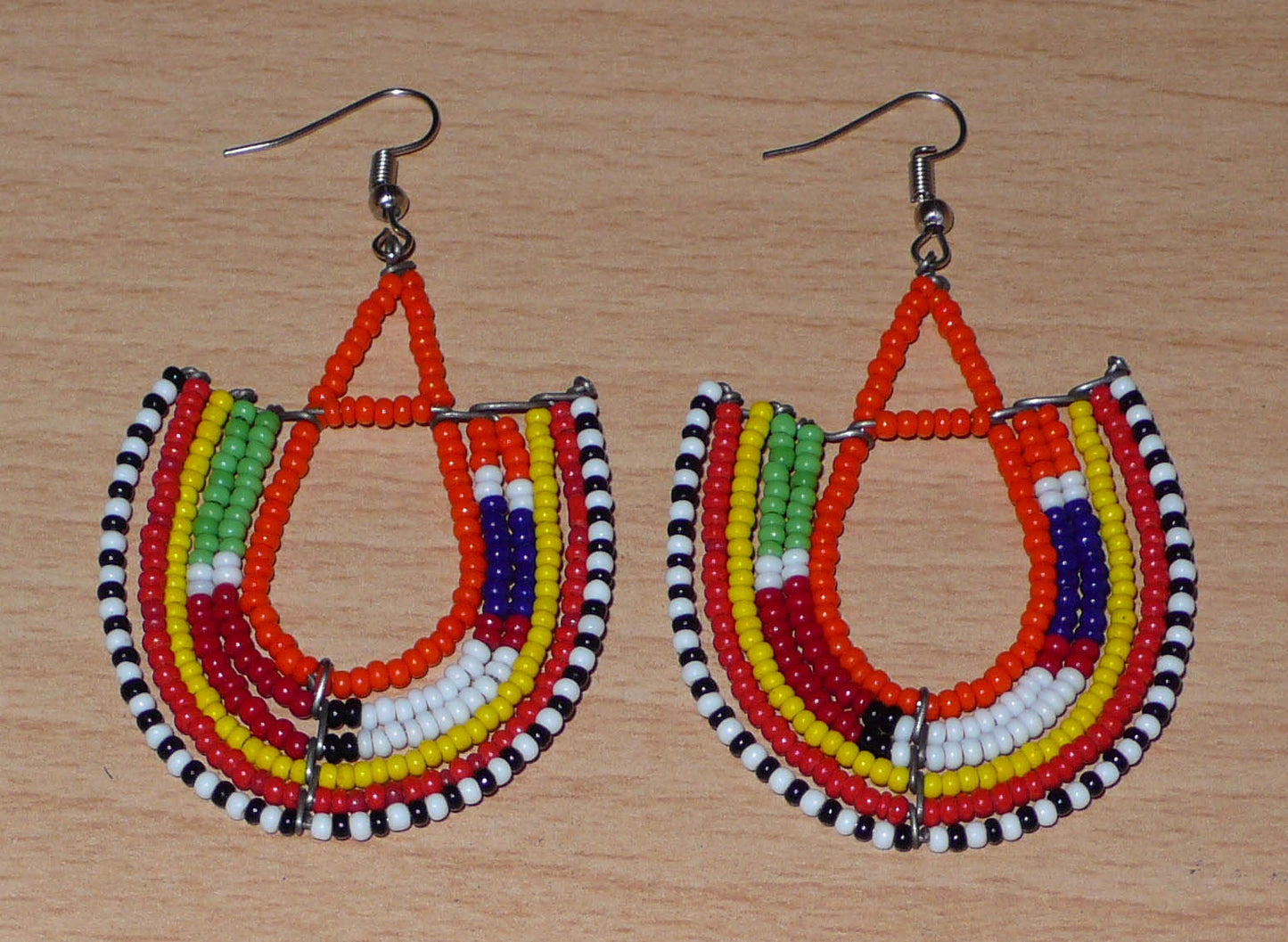 Boucles d'oreilles africaines traditionnelles aux couleurs et motifs ethniques Massai caractéristiques de l'artisanat kenyan. Elles se composent de perles de rocaille orange et multicolores fixées sur des structures en fils de fer en forme d'ancres. Ces boucles d'oreilles tribales mesurent 7.5 cm de long et 4.5 cm de large ; elles se portent avec des crochets en acier inoxydable sur des oreilles percées.