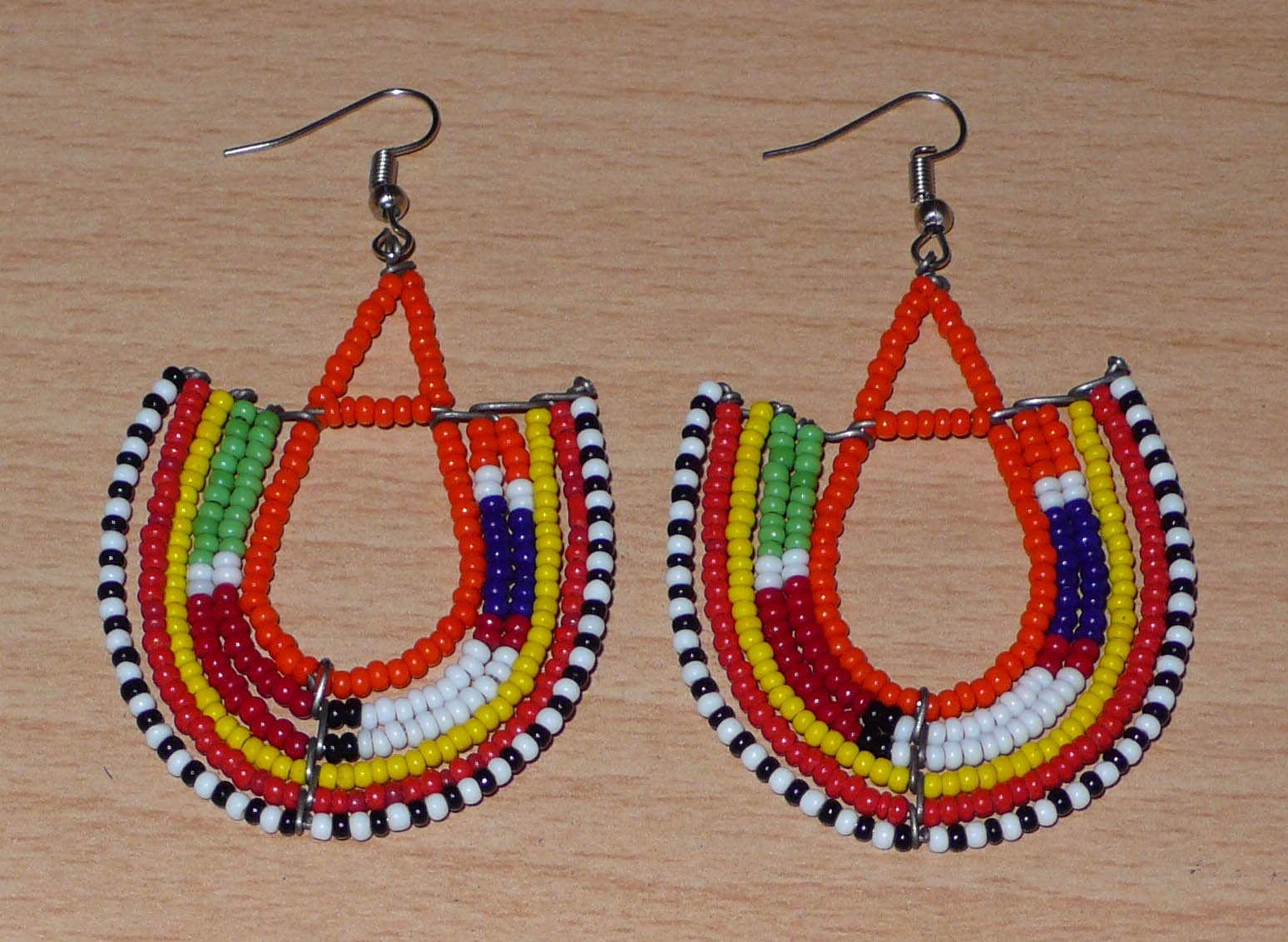 Boucles d'oreilles africaines traditionnelles aux couleurs et motifs ethniques Massai caractéristiques de l'artisanat kenyan. Elles se composent de perles de rocaille orange et multicolores fixées sur des structures en fils de fer en forme d'ancres. Ces boucles d'oreilles tribales mesurent 7.5 cm de long et 4.5 cm de large ; elles se portent avec des crochets en acier inoxydable sur des oreilles percées.