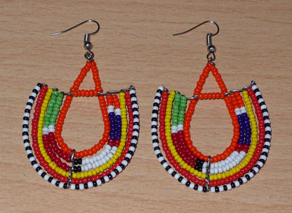Boucles d'oreilles africaines traditionnelles aux couleurs et motifs ethniques Massai caractéristiques de l'artisanat kenyan. Elles se composent de perles de rocaille orange et multicolores fixées sur des structures en fils de fer en forme d'ancres. Ces boucles d'oreilles tribales mesurent 7.5 cm de long et 4.5 cm de large ; elles se portent avec des crochets en acier inoxydable sur des oreilles percées.