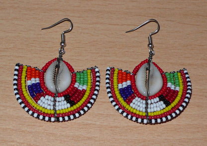 Ajoutez une touche exotique à votre style avec cette jolie paire de boucles d'oreilles africaines issues de l'artisanat traditionnel Massai. Ces boucles d'oreilles ethniques chic se composent de perles de rocaille rouges et multicolores et de coquillages cauris. De taille moyenne, elles mesurent 6 cm de long et 4.5 cm de large, et se portent avec des crochets en acier inoxydable sur des oreilles percées.