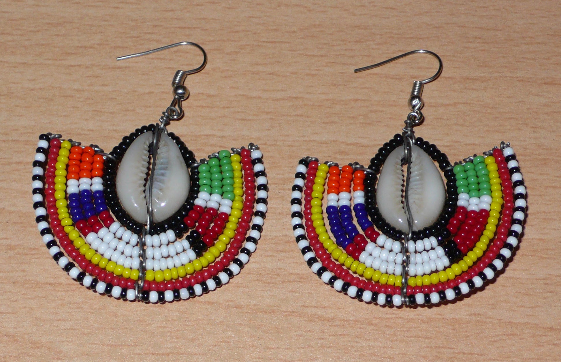 Apportez une note de couleurs à votre look avec cette paire de boucles d'oreilles africaines au style exotique chic issues de l'artisanat traditionnel Massai. Confectionnées à la main au Kenya, ces boucles d'oreilles se composent de perles de rocaille noires et multicolores et de coquillages cauris. De taille moyenne, elles mesurent 6 cm de long et 4.5 cm de large, et se portent avec des crochets en acier inoxydable sur des oreilles percées.