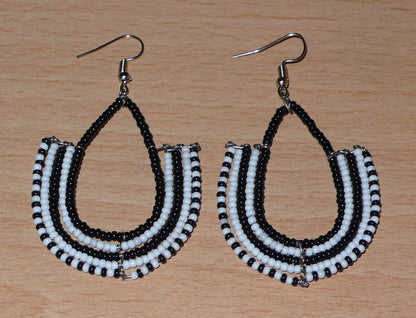 Boucles d'oreilles kenyanes Massai bicolores en forme d'ancres composées de perles de rocaille noires et blanches glissées sur des structures géométriques en fils de fer. De taille moyenne, ces boucles d'oreilles aux accents ethniques afro chic mesurent 7 cm de long et 4 cm de large ; elles se portent avec des crochets en acier inoxydable sur des oreilles percées.
