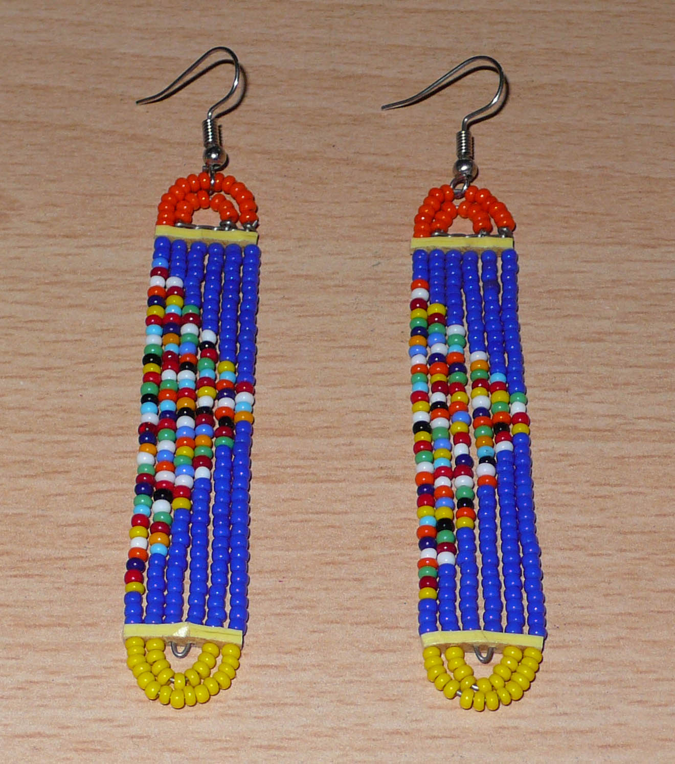 Longues boucles d'oreilles kenyanes en perles de rocaille bleues, orange, jaunes et multicolores glissées sur six rangées de fils de fer montés en forme de rectangles aux bouts arrondis. Elles mesurent 9,5 cm de long et 1,5 cm de large et se portent sur des oreilles percées.