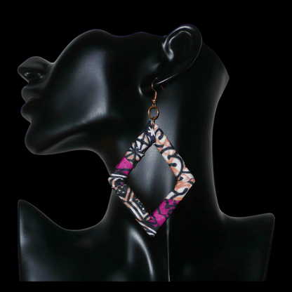 Longues boucles d'oreilles africaines en forme de losanges composées de tissu africain rose-violet, avec des touches de noir, de blanc et de beige. Elles mesurent 11 cm de long et 6 cm de large et se portent sur des oreilles percées.