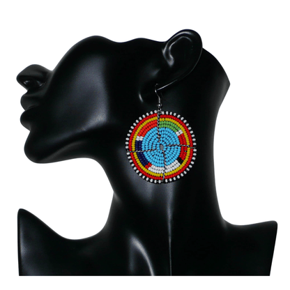 Boucles d'oreilles africaines rondes à motifs ethniques Massai composées de perles de rocaille bleues et multicolores. Elles mesurent 6.5 cm de long et 5 cm de large, et se portent sur des oreilles percées.