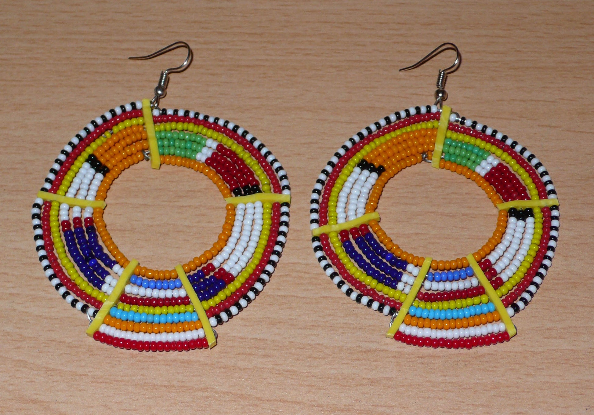 Boucles d'oreilles africaines tribales surdimensionnées, fabriquées au Kenya dans la plus pure tradition artisanale Massai. Elles se composent de perles de rocaille orange et multicolores tissées sur des structures circulaires en fils de fer fixés à des charnières en plastique jaune. Ces longues et larges boucles d'oreilles ethniques mesurent 8.5 cm de long et 6 cm de large ; elles se portent avec des crochets en acier inoxydable sur des oreilles percées.