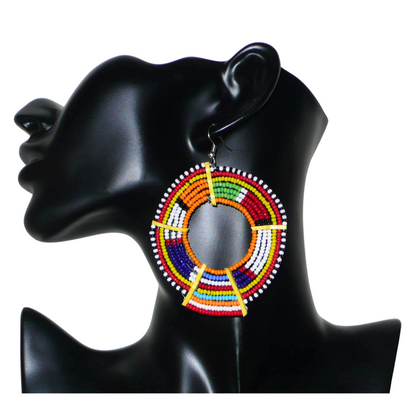 Boucles d'oreilles africaines tribales surdimensionnées, fabriquées au Kenya dans la plus pure tradition artisanale Massai. Elles se composent de perles de rocaille orange et multicolores tissées sur des structures circulaires en fils de fer fixés à des charnières en plastique jaune. Ces longues et larges boucles d'oreilles ethniques mesurent 8.5 cm de long et 6 cm de large ; elles se portent avec des crochets en acier inoxydable sur des oreilles percées.