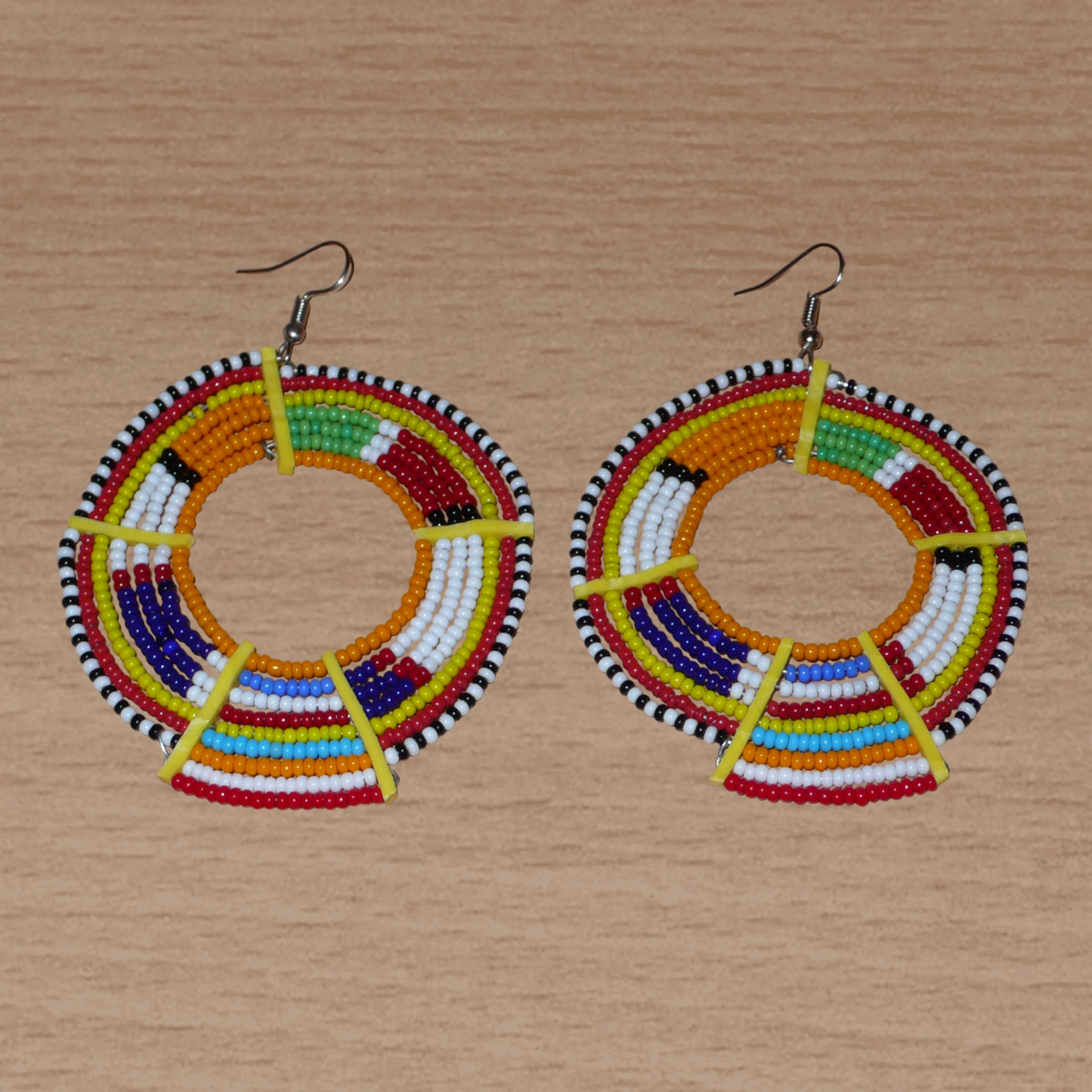Boucles d'oreilles africaines tribales surdimensionnées, fabriquées au Kenya dans la plus pure tradition artisanale Massai. Elles se composent de perles de rocaille orange et multicolores tissées sur des structures circulaires en fils de fer fixés à des charnières en plastique jaune. Ces longues et larges boucles d'oreilles ethniques mesurent 8.5 cm de long et 6 cm de large ; elles se portent avec des crochets en acier inoxydable sur des oreilles percées.