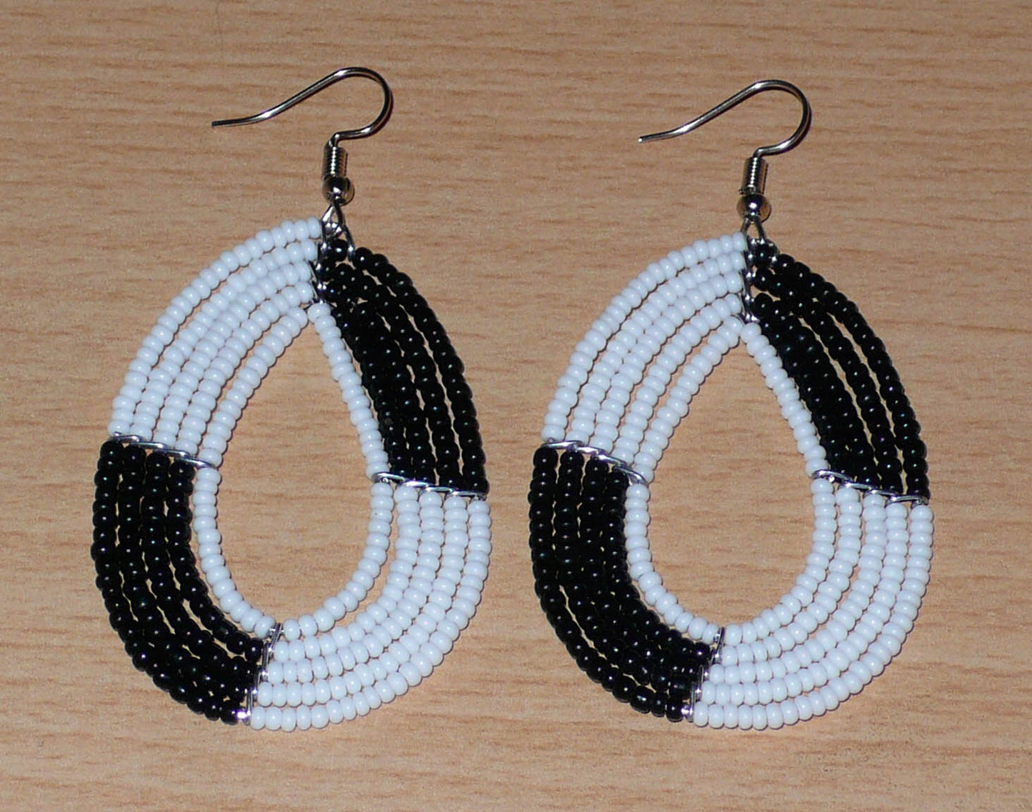 Boucles d'oreilles bicolores noires et blanches en forme de gouttes faites de perles de rocaille enfilées sur cinq rangées de fils. Elles mesurent 7,5 cm de long et 3 cm de large et se portent sur des oreilles percées.