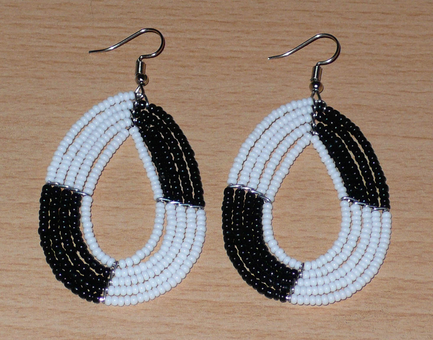 Boucles d'oreilles bicolores noires et blanches en forme de gouttes faites de perles de rocaille enfilées sur cinq rangées de fils. Elles mesurent 7,5 cm de long et 3 cm de large et se portent sur des oreilles percées.