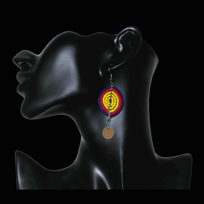 Boucles d'oreilles africaines en forme de boucliers Massai composées de perles de rocaille jaunes, rouges et bleu foncé ; elles se prolongent d'un disque métallique. Ces boucles d'oreilles mesurent 7 cm de long et 2,5 cm de large ; elles se portent sur des oreilles percées.
