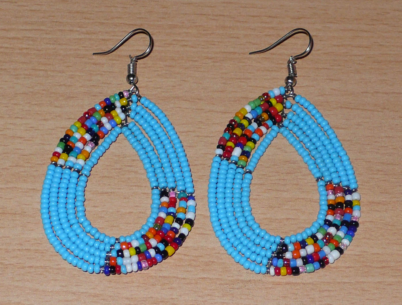 Pendientes étnicos de varias hileras compuestos por cuentas azul cielo y multicolores deslizadas sobre cinco hileras de alambre montado en forma de gotas. De tamaño mediano, estos aretes kenianos con detalles bohemios miden 7,5 cm de largo y 4 cm de ancho; Se llevan con ganchos de acero inoxidable.