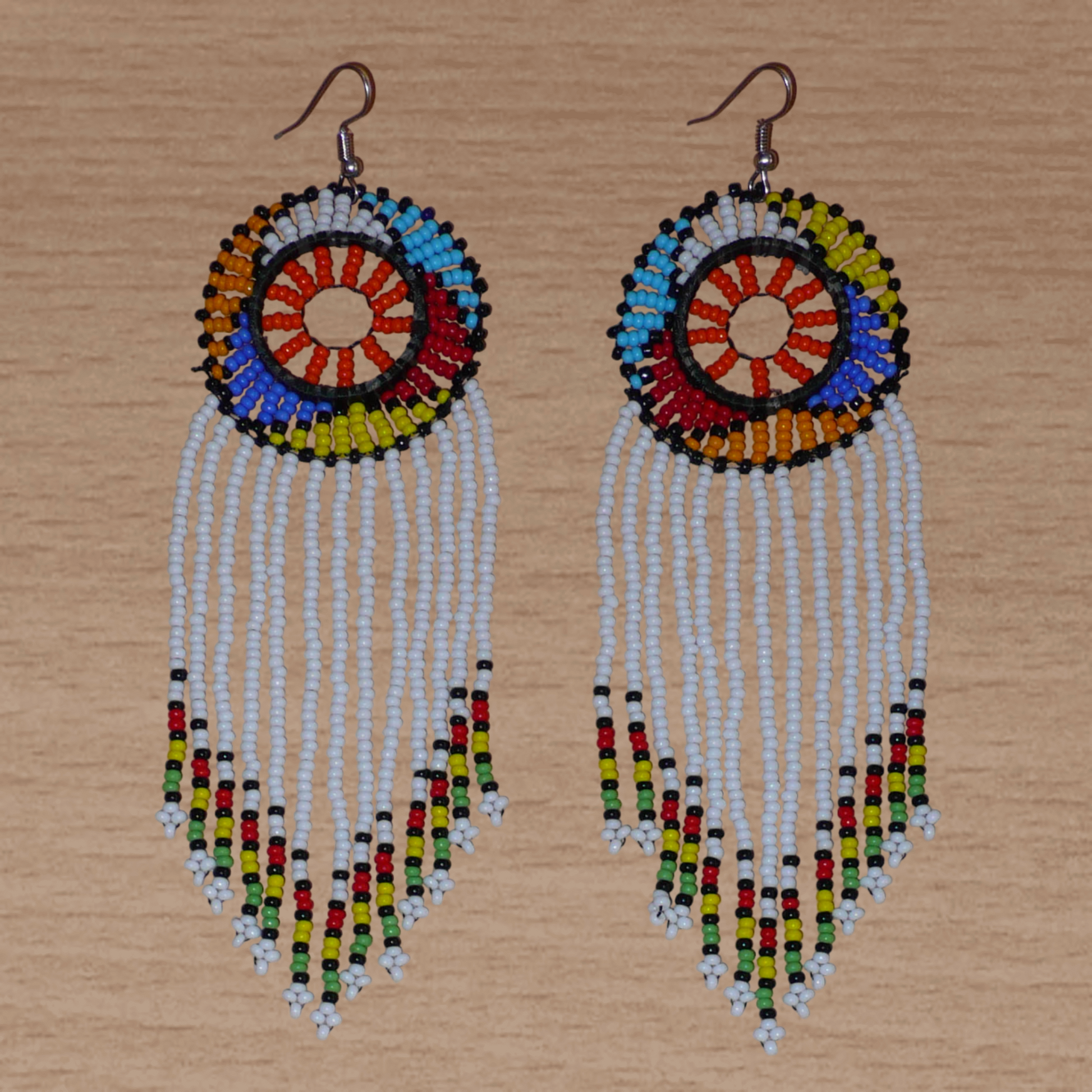 Longues boucles d'oreilles sud-africaines à franges et motifs traditionnels zoulous composées de perles de rocaille blanches, orange et multicolores. Elles mesurent 14,5 cm de long et 4,5 cm de large, et se portent avec des crochets en acier inoxydable sur des oreilles percées.