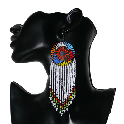 Longues boucles d'oreilles sud-africaines à franges et motifs traditionnels zoulous composées de perles de rocaille blanches, orange et multicolores. Elles mesurent 14,5 cm de long et 4,5 cm de large, et se portent avec des crochets en acier inoxydable sur des oreilles percées.