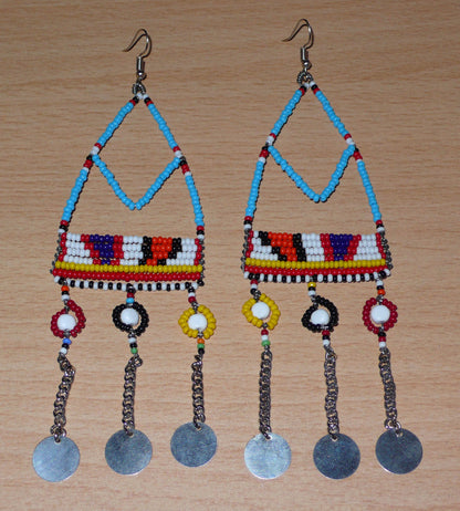 Longues boucles d'oreilles africaines Massai bleues et multicolores composées de perles de rocaille bleues et multicolores et de perles en plastique blanc glissées sur des structures géométriques en fils de fer ; elles se prolongent de chaînettes décorées de disques métalliques. Surdimensionnées, elles mesurent 15.5 cm de long et se portent sur des oreilles percées.