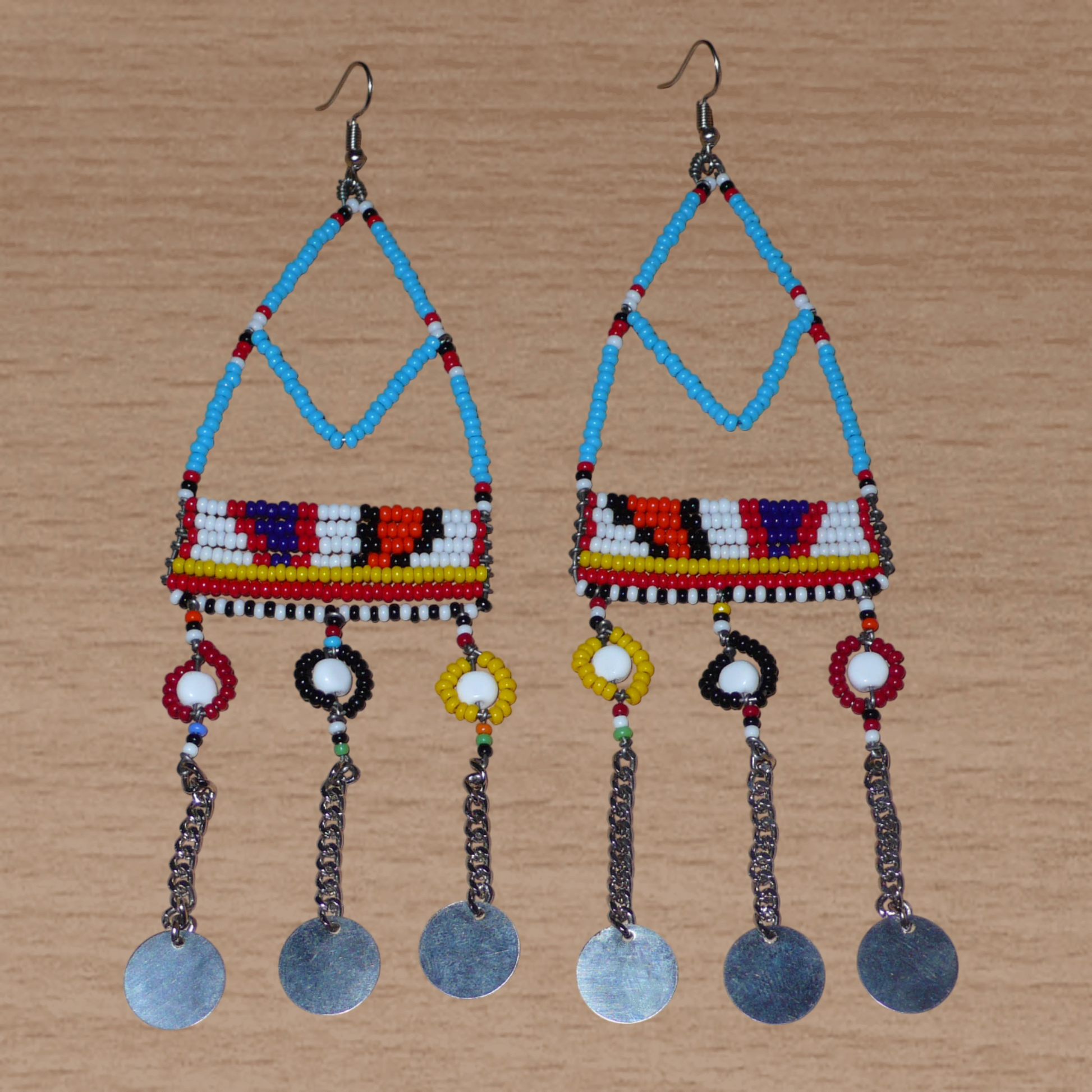 Longues boucles d'oreilles africaines Massai bleues et multicolores composées de perles de rocaille bleues et multicolores et de perles en plastique blanc glissées sur des structures géométriques en fils de fer ; elles se prolongent de chaînettes décorées de disques métalliques. Surdimensionnées, elles mesurent 15.5 cm de long et se portent sur des oreilles percées.