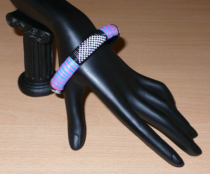 Bracelet africain quatre couleurs à motifs ethniques tissés avec des fines bandes de plastique recyclé noir, blanc, rose et bleu.  Le bracelet a une circonférence de 20 cm, un diamètre de 6,5 cm et une épaisseur de 1,3 cm.  Il peut être porté par une femme ou par un homme.