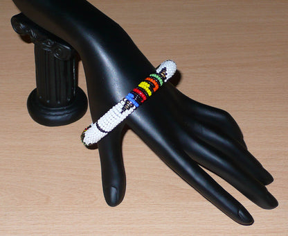 Bracelet africain tubulaire à motifs traditionnels zoulous composé de perles de rocaille blanches et multicolores ; glissées sur du fil de nylon, les perles sont ensuite enroulées autour d'un tube en plastique. Ce bracelet a une circonférence de 21 cm, un diamètre de 6.5 cm, et une épaisseur de 1 cm.