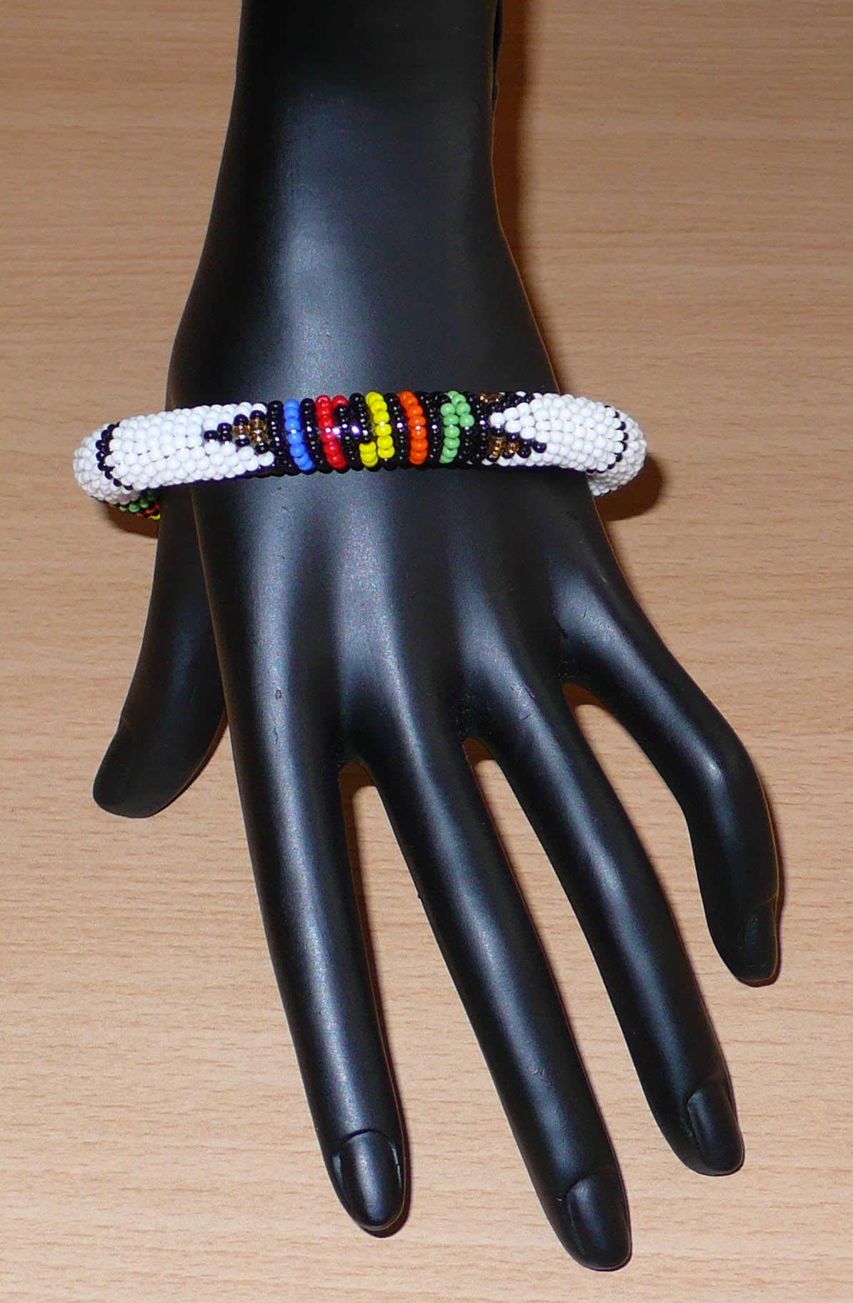 Bracelet africain tubulaire à motifs traditionnels zoulous composé de perles de rocaille blanches et multicolores ; glissées sur du fil de nylon, les perles sont ensuite enroulées autour d'un tube en plastique. Ce bracelet a une circonférence de 21 cm, un diamètre de 6.5 cm, et une épaisseur de 1 cm.
