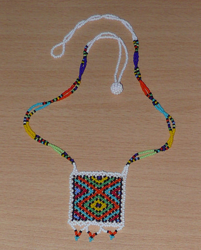 Collier africain orné d'un pendentif carré à motifs ethniques zoulous décoré d'une petite frise perlée. Il se compose d'un tissage de perles de rocaille blanches et multicolores et s'attache avec un fermoir à boule.