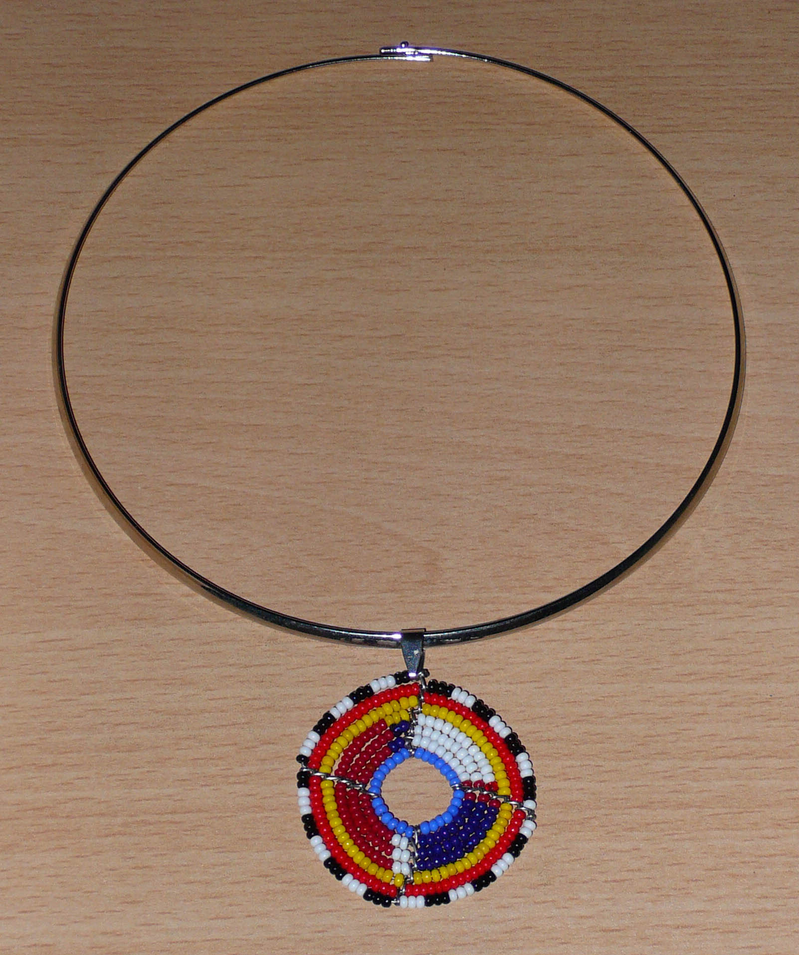 Collier africain composé d'un ras de cou en laiton orné d'un petit médaillon fait d'un tissage de perles de rocaille. Il mesure 18,5 cm de long, la circonférence du ras de cou est de 42,5 cm et son diamètre de 13,5 cm ; le médaillon mesure 4,5 cm de long et de large.