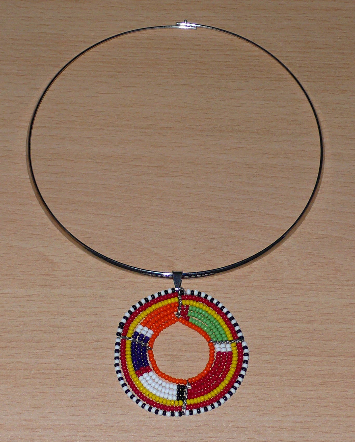 Alliance de tradition kenyane et de modernité, ce collier se compose d'un ras de cou en laiton agrémenté d'un médaillon perlé aux couleurs et motifs ethniques traditionnels Massai fait d'un tissage de perles de rocaille multicolores.