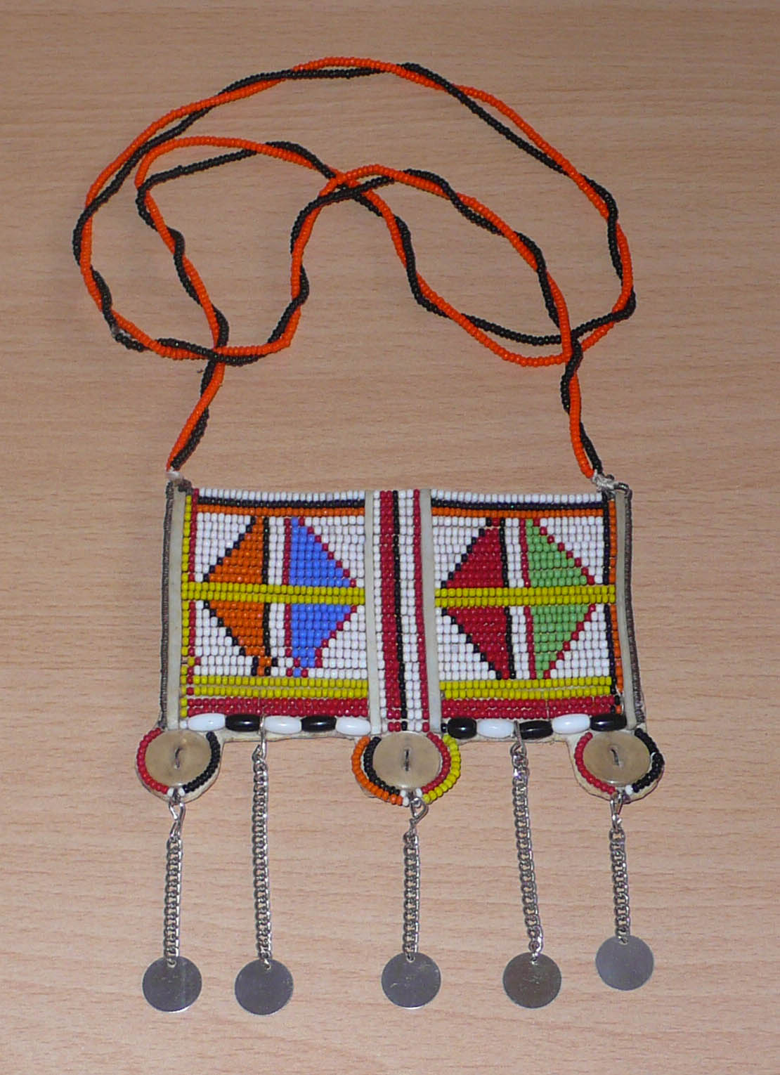 Long collier africain Massai traditionnel de style sautoir orné d'un large pendentif à motifs tribaux fait de perles brodées et de boutons nacrés ; il se prolonge de chaînettes décorées de pastilles métalliques.