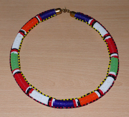 Collier africain tubulaire à motifs traditionnels Massai composé de perles de rocaille rouges, vertes, blanches, bleu foncé et orange. Il mesure 22 cm de long, la circonférence autour du cou est de 56 cm et son épaisseur de 1,5 cm. Timeless Fineries