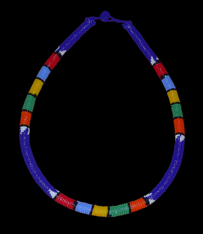 Collier sud-africain tubulaire bleu et multicolore. Il se compose d'un tube de plastique autour duquel sont enroulées des petites perles de rocaille bleues alternées avec des motifs ethniques traditionnels zoulous rouges, bleu ciel, jaunes, verts et orange. Le collier mesure 22 cm de long (de la nuque jusqu'à la base du collier), la longueur du tour de cou du collier est 56 cm, et son épaisseur est de 1 cm. Il s'attache avec un fermoir à boule. Collier photographié sur un fond noir. Timeless Fineries