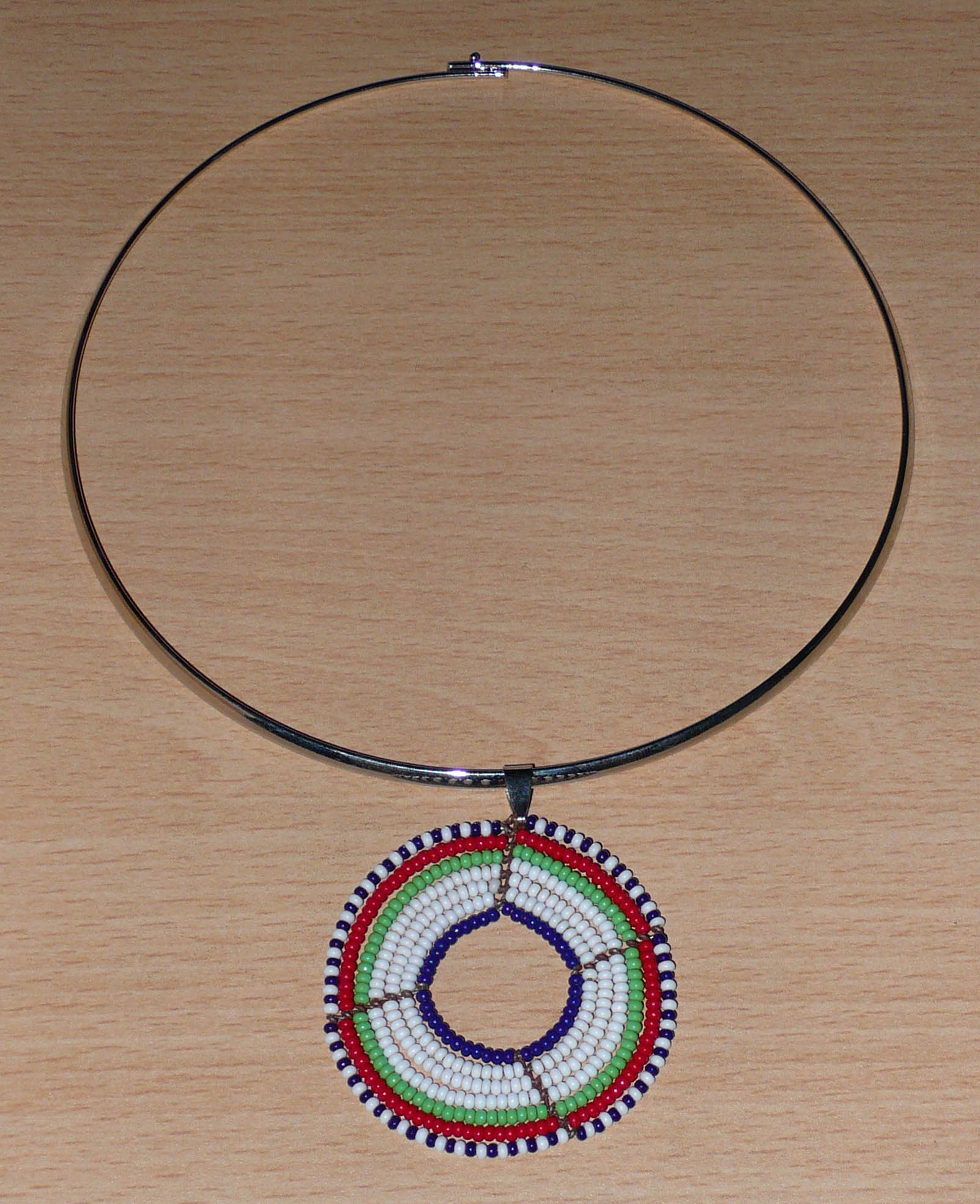 Collier ethnique contemporain composé d'un ras de cou en laiton orné d'un médaillon Massai fait de perles de rocaille blanches, vertes, rouges, bleu marine et noires.