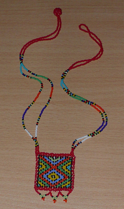 Collier africain en perles de rocaille rouges et multicolores agrémenté d'un pendentif carré à motifs ethniques. Il mesure 27,5 cm de long (en partant de la nuque jusqu'à la base du pendentif), le pendentif mesure 5,5 cm sur 4,5 cm ; le collier s'attache avec un fermoir à boule. Timeless Fineries