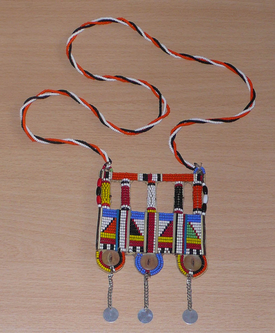 Authentique bijou africain Massai, ce long collier de style sautoir est orné d'un large pendentif à motifs ethniques fait de perles de rocaille multicolores et de boutons nacrés brodés sur un morceau de plastique recyclé ; ils se prolonge de chaînettes décorées de pastilles métalliques.