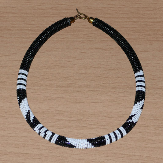 Collier africain tubulaire noir à motifs tribaux zoulous blancs composé de perles de rocaille tissées et enroulées autour d'un tube. Il s'attache avec un crochet.