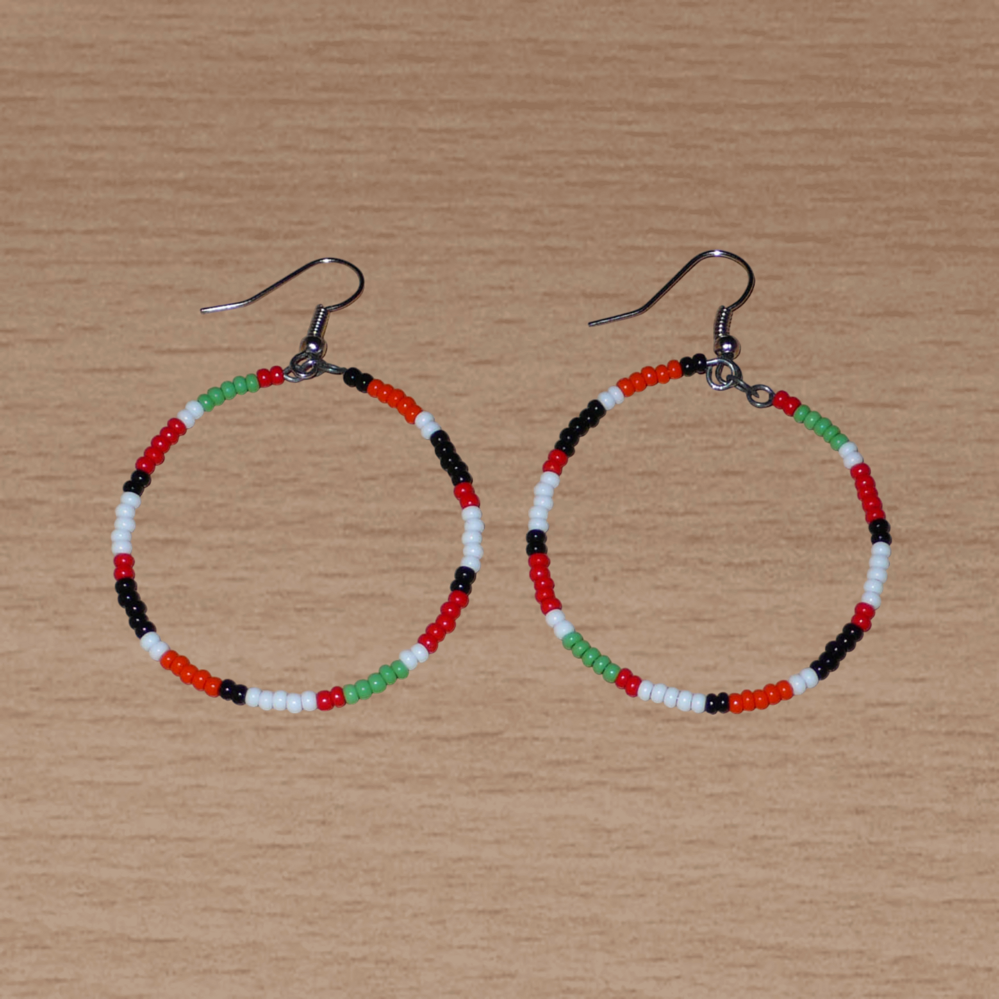 Créoles aux couleurs ethniques Massai faites de perles de rocaille rouges, vertes, noires, blanches et orange glissées sur un brin de fils de fer monté en cercle. Elles mesurent 6 cm de long et 4 cm de large, et se portent avec des crochets en acier inoxydable sur des oreilles percées.