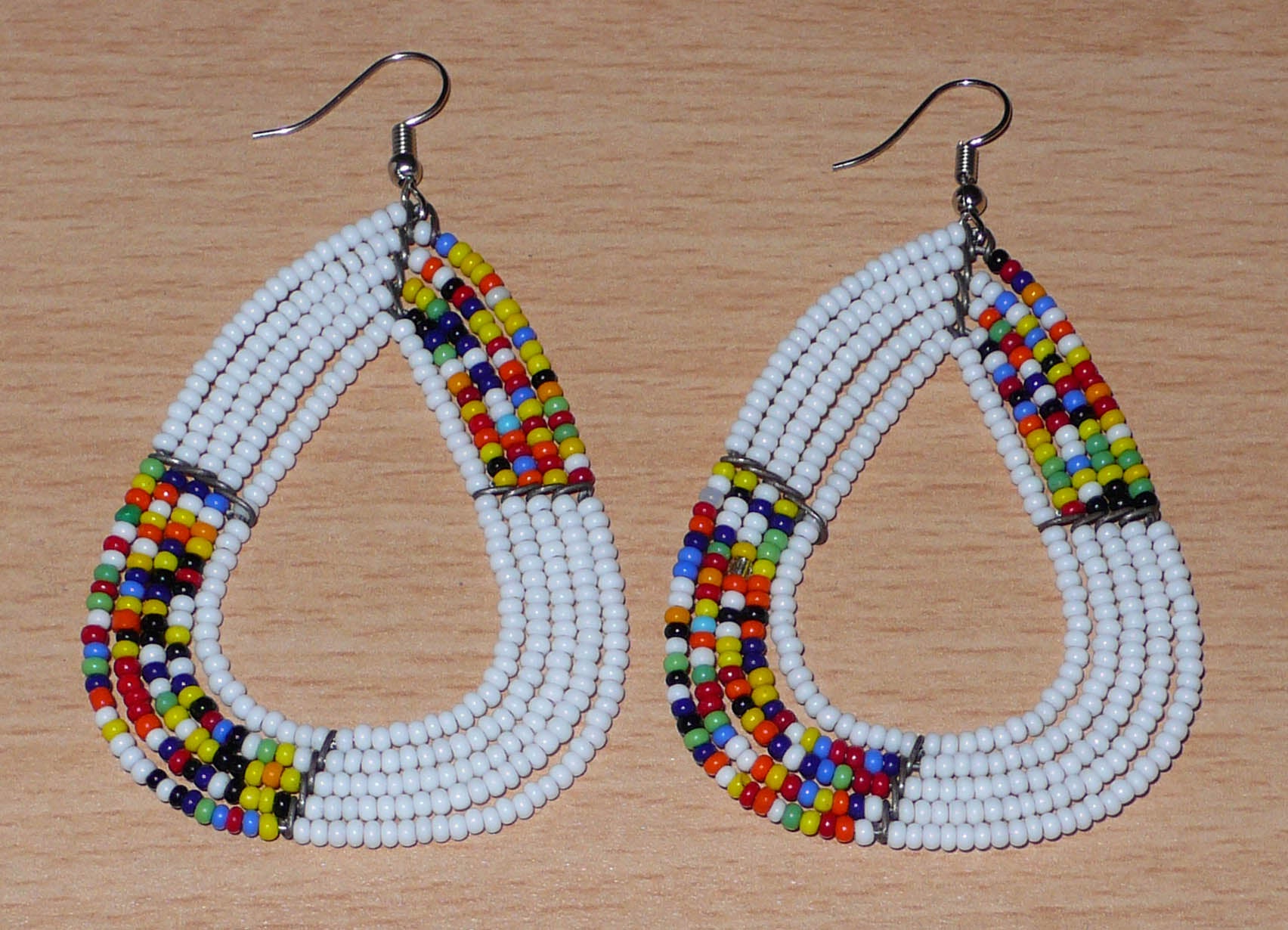 Longues et larges boucles d'oreilles kenyanes composées de perles de rocaille blanches et multicolores glissées sur cinq rangées de fils de fer montés en forme de gouttes légèrement triangulaires. Elles mesurent 8 cm de long et 5 cm de large et se portent avec des crochets en acier inoxydable sur des oreilles percées.