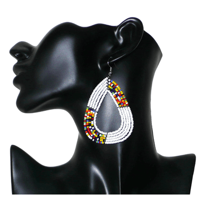Longues et larges boucles d'oreilles kenyanes composées de perles de rocaille blanches et multicolores glissées sur cinq rangées de fils de fer montés en forme de gouttes légèrement triangulaires. Elles mesurent 8 cm de long et 5 cm de large et se portent avec des crochets en acier inoxydable sur des oreilles percées.